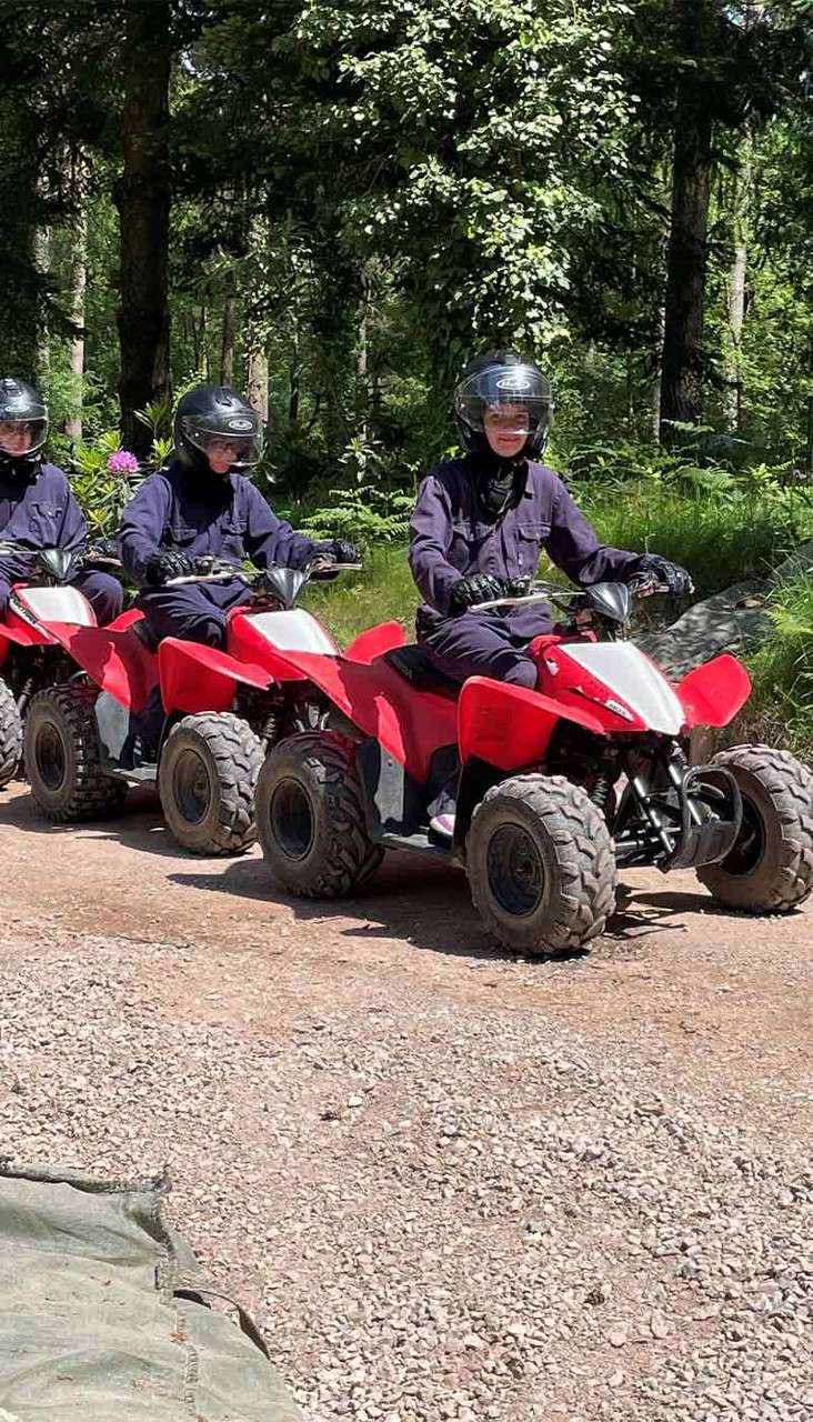 Three helmeted riders drive red quad bikes in a line along a gravel trail, surrounded by green forest trees and ferns in sunlight.