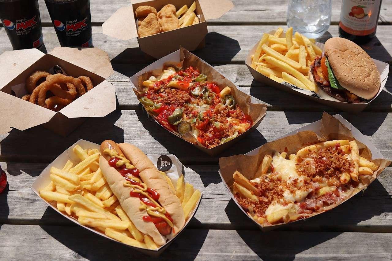 Trays of fast food sit arranged on a sunlit picnic table: hot dog with ketchup and mustard, burger, fries, onion rings, nuggets, loaded fries, and drinks; two cups read pepsi MAX.