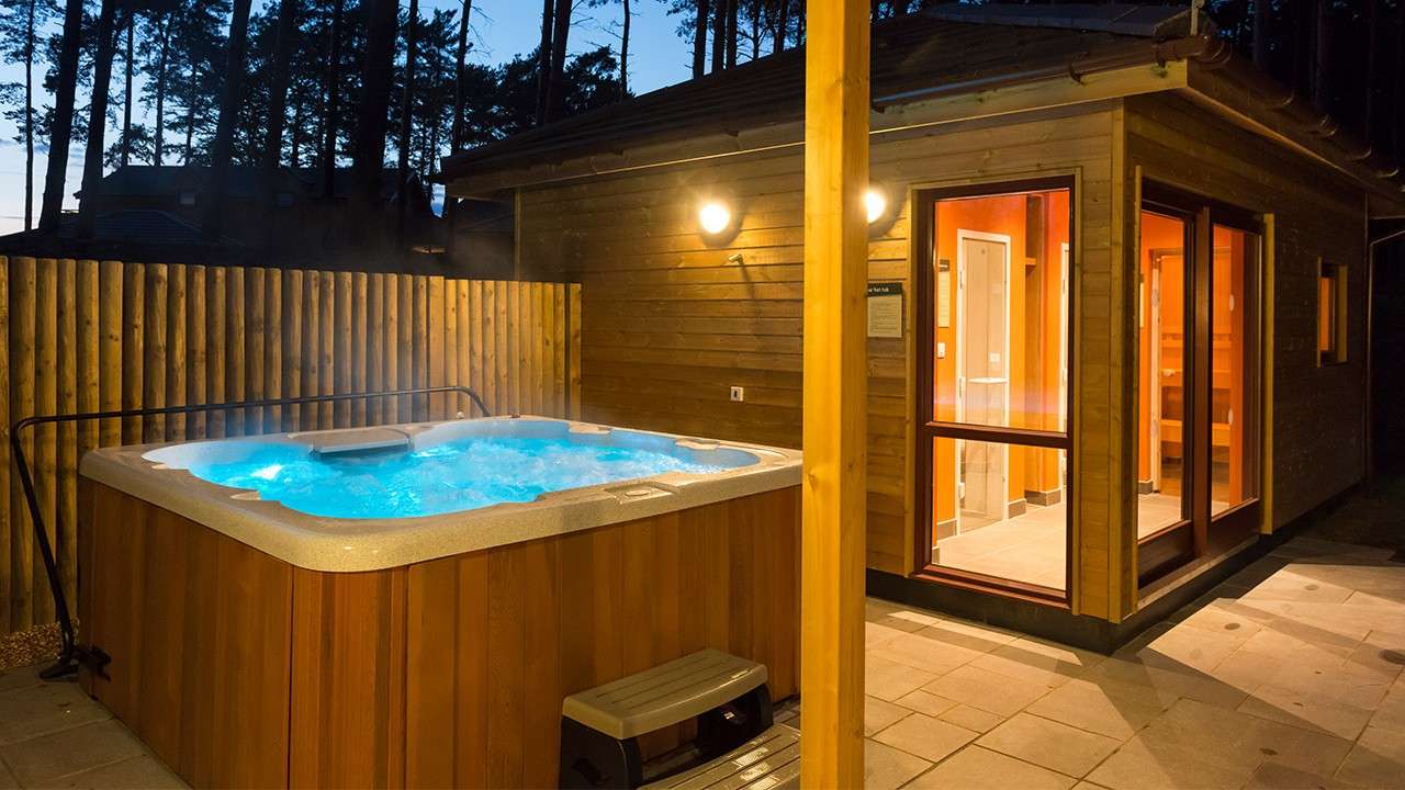 Hot tub bubbles and steams, glowing blue, on a fenced patio. Next to it, a lit wooden cabin with large windows stands under dusk sky, pine trees silhouetted around.