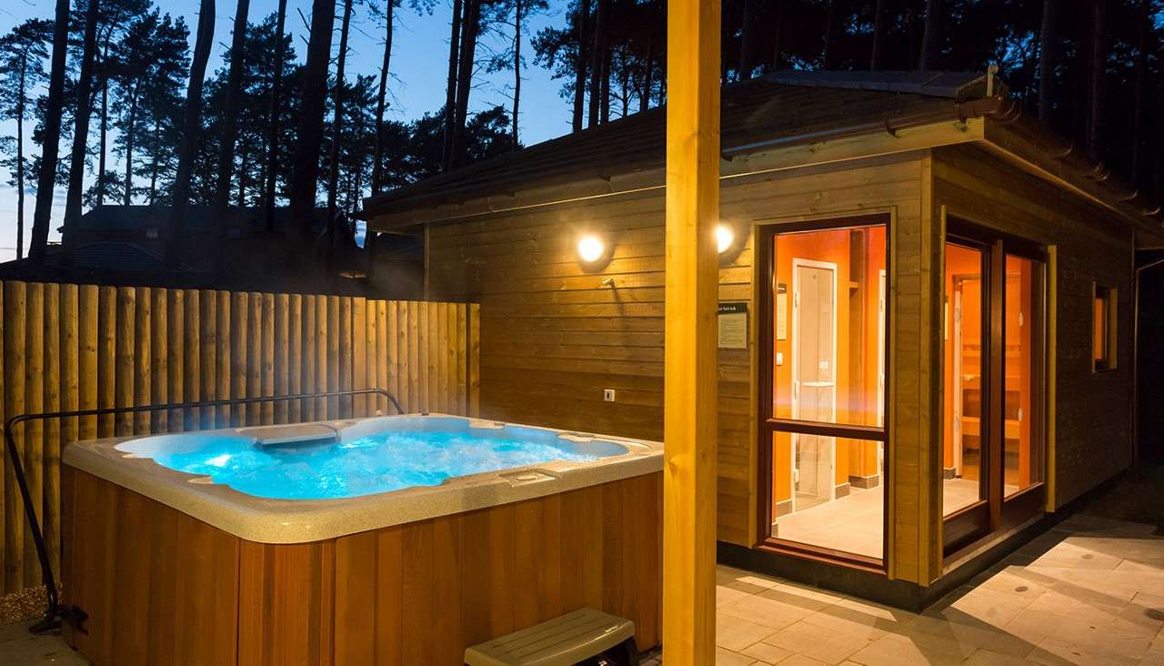Hot tub bubbles and steams, its blue-lit water churning. Set on a patio beside a warmly lit wooden cabin, enclosed by a fence, with tall pine trees silhouetted at dusk.