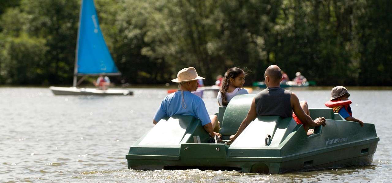 Green pedal boat carries four people, moving across a calm lake; nearby sailboats glide and trees line the shore. Text on boat reads: "pioner tamar".