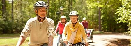 Cyclists ride along a sunlit forest path, wearing helmets and casual clothing. Two adults lead while others follow, including a child in a bike trailer. Trees and light surround.