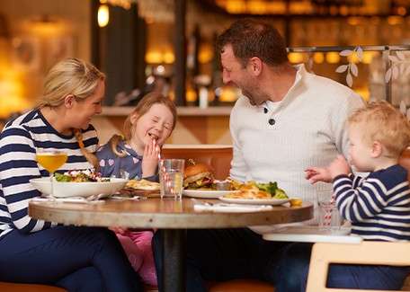 Parents and two young children laugh while eating at a round restaurant table with salad, burger, fries, and drinks, surrounded by warm indoor lighting and softly blurred decor.