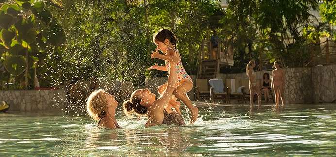 Child in a polka-dot swimsuit is lifted playfully by two adults, splashing water, in a sunlit outdoor pool surrounded by trees, lounge chairs, and other swimmers in the background.
