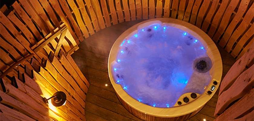 Round hot tub bubbles with steam, blue LED lights glowing around its rim. Viewed from above, it sits on a wooden deck, enclosed by fencing and a wall lamp.