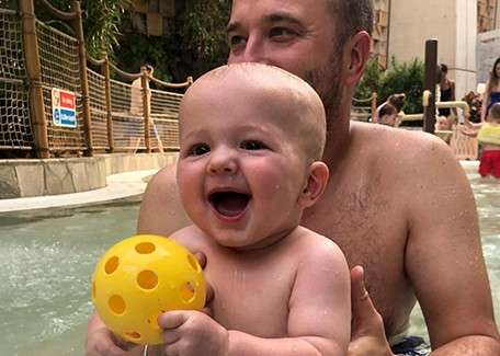Baby laughs holding a yellow perforated ball; an adult supports from behind. They sit in shallow pool water at an indoor waterpark with rope fencing and other swimmers in background.