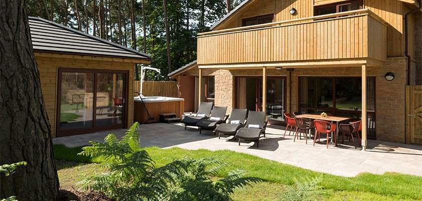 Patio furniture sits under a wooden balcony; lounge chairs and a dining table arranged on a stone terrace; a hot tub sits nearby; surrounded by grass lawn and forested trees.
