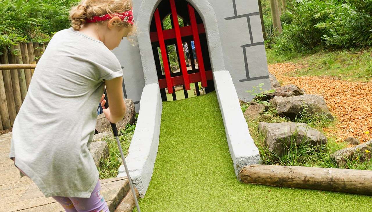 Child putts a mini-golf ball toward a castle-themed tunnel gate on a green ramp; wooden barriers, rocks, and trees surround the outdoor course.
