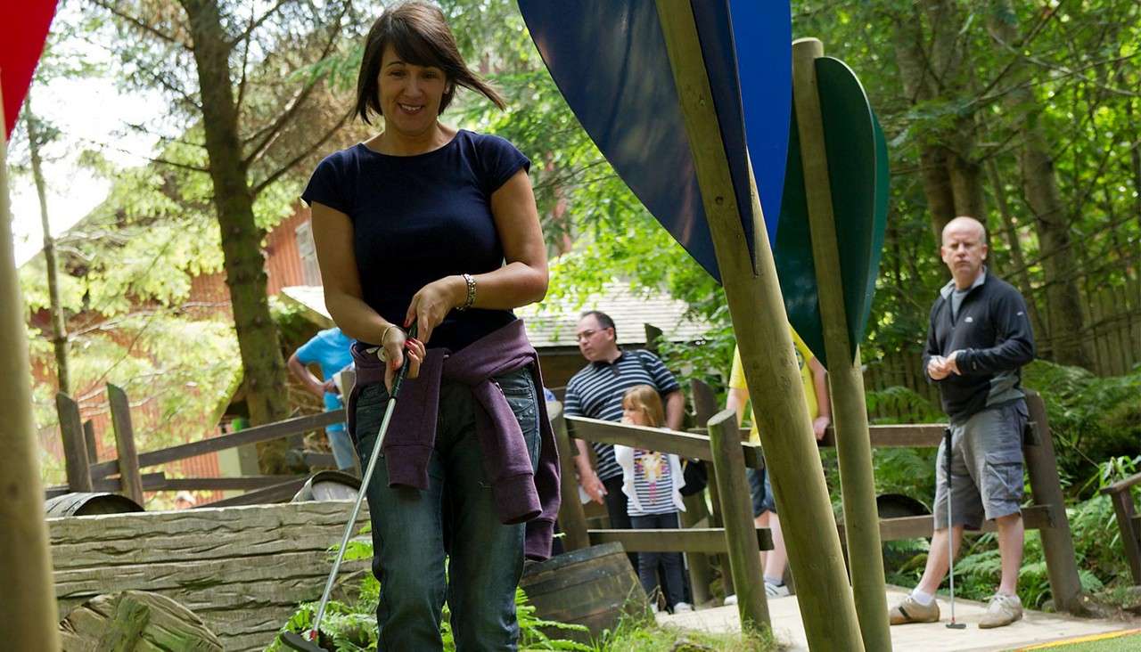 Woman putts a golf ball with a putter, smiling and focused, while other players wait behind wooden rails on a forested mini-golf course with colorful obstacles and barrels.
