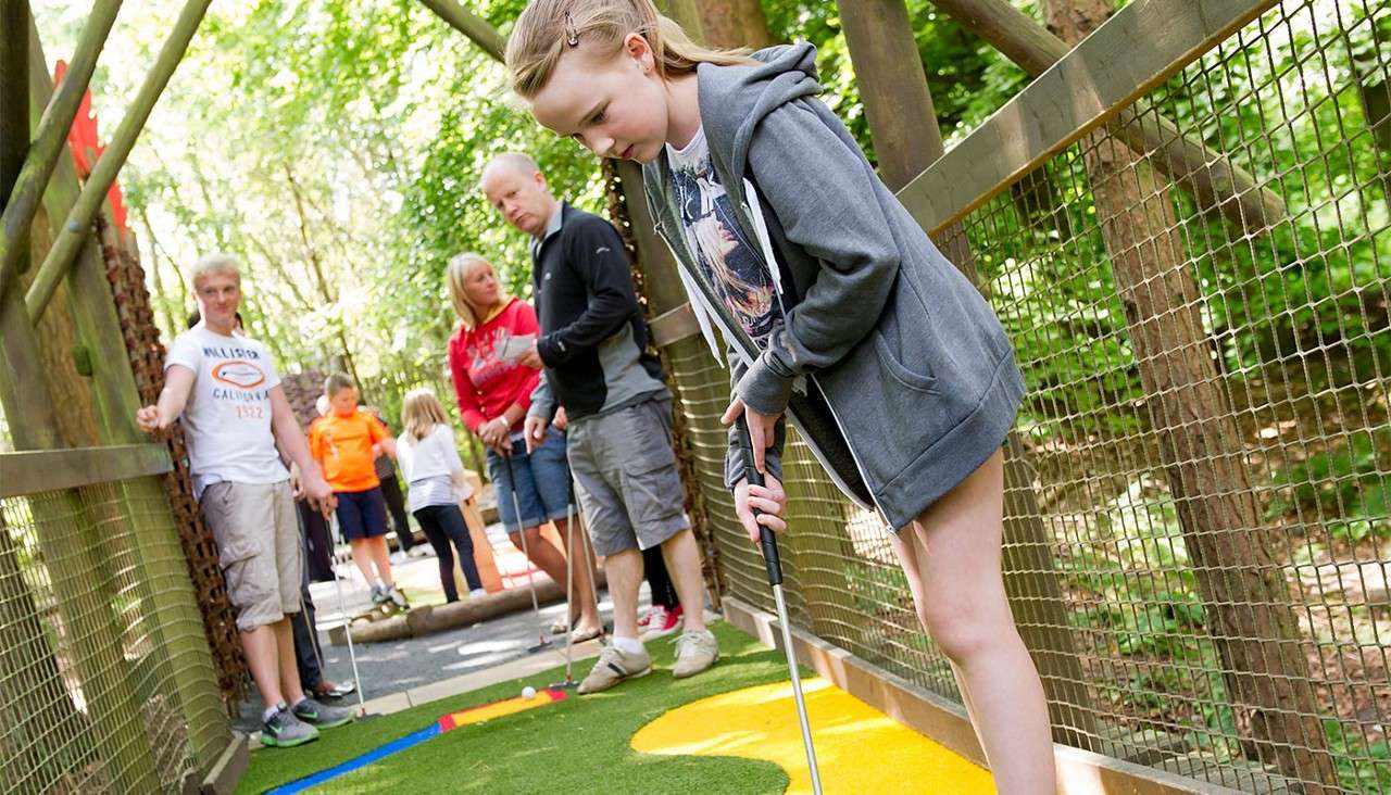 Girl golfer lines up a putt, gripping a club, on a colorful mini-golf lane. Families watch nearby within a wooded, netted walkway course. Text: "HOLLISTER CALIFORNIA 1922".