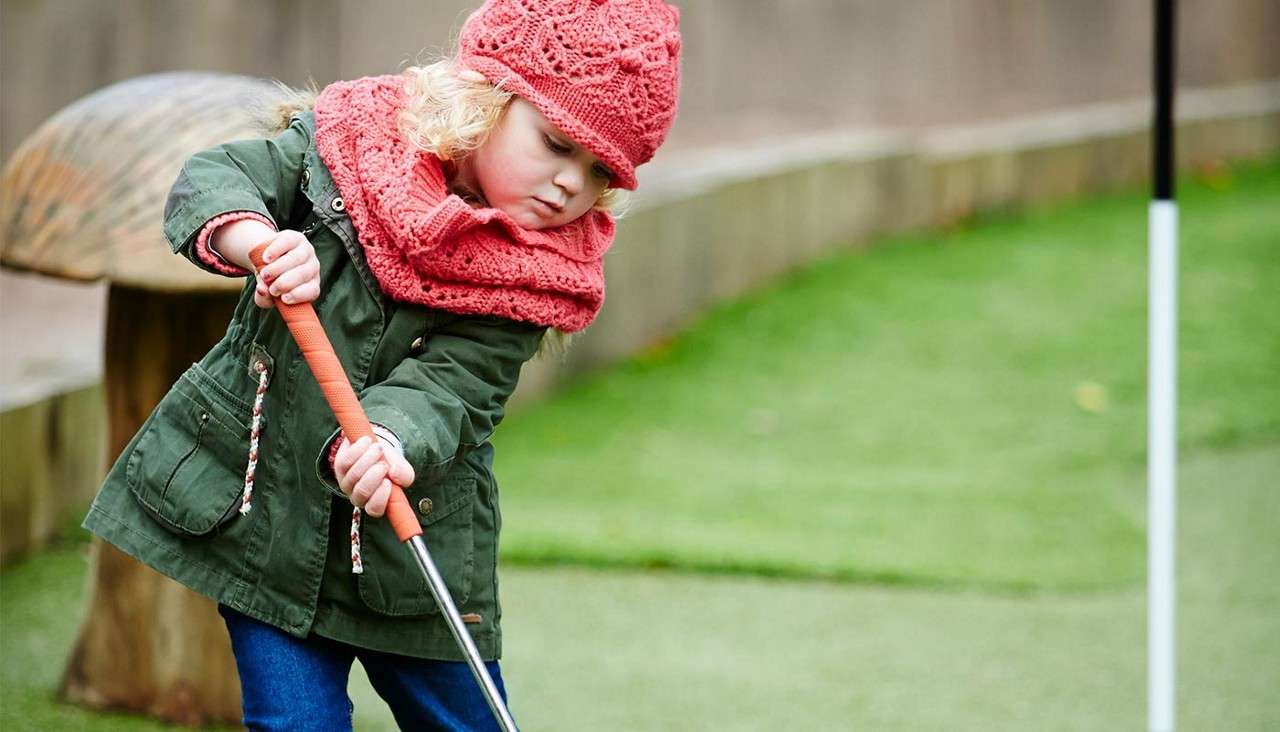Child in green coat and pink knitted hat and scarf swings a mini-golf putter on an outdoor putting green, concentrating near a hole marker and low wooden edging.
