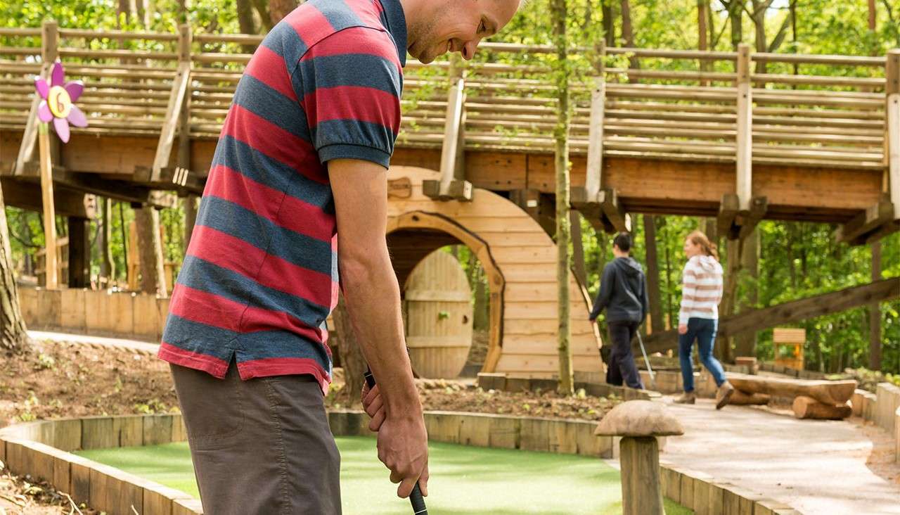 Man putts a mini-golf ball; others walk nearby; wooden bridge and tunnel structures stand in a forest-themed course. A flower-shaped hole sign reads “6.”