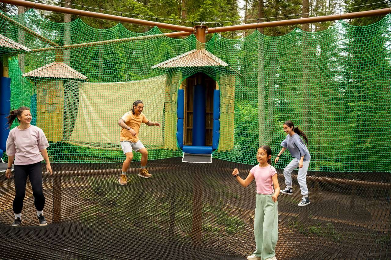 Four people bounce on a large suspended net, playing inside green mesh walls with a small hut and slide doorway, surrounded by tall forest trees and wooden supports.