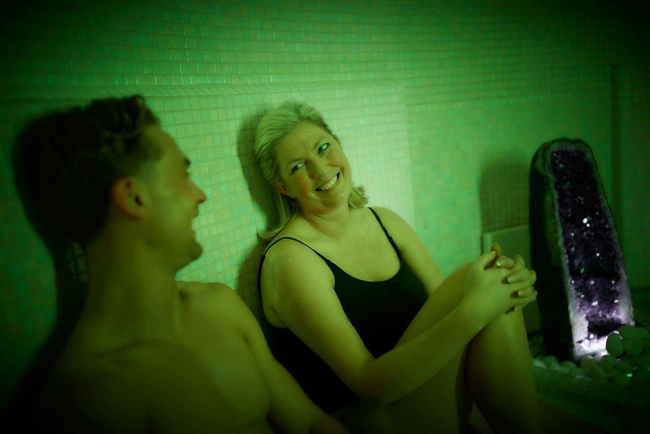 Two adults sit relaxing, smiling and chatting, in swimsuits. They lean against tiled walls under green light; a lit amethyst geode water feature stands beside them among white pebbles.