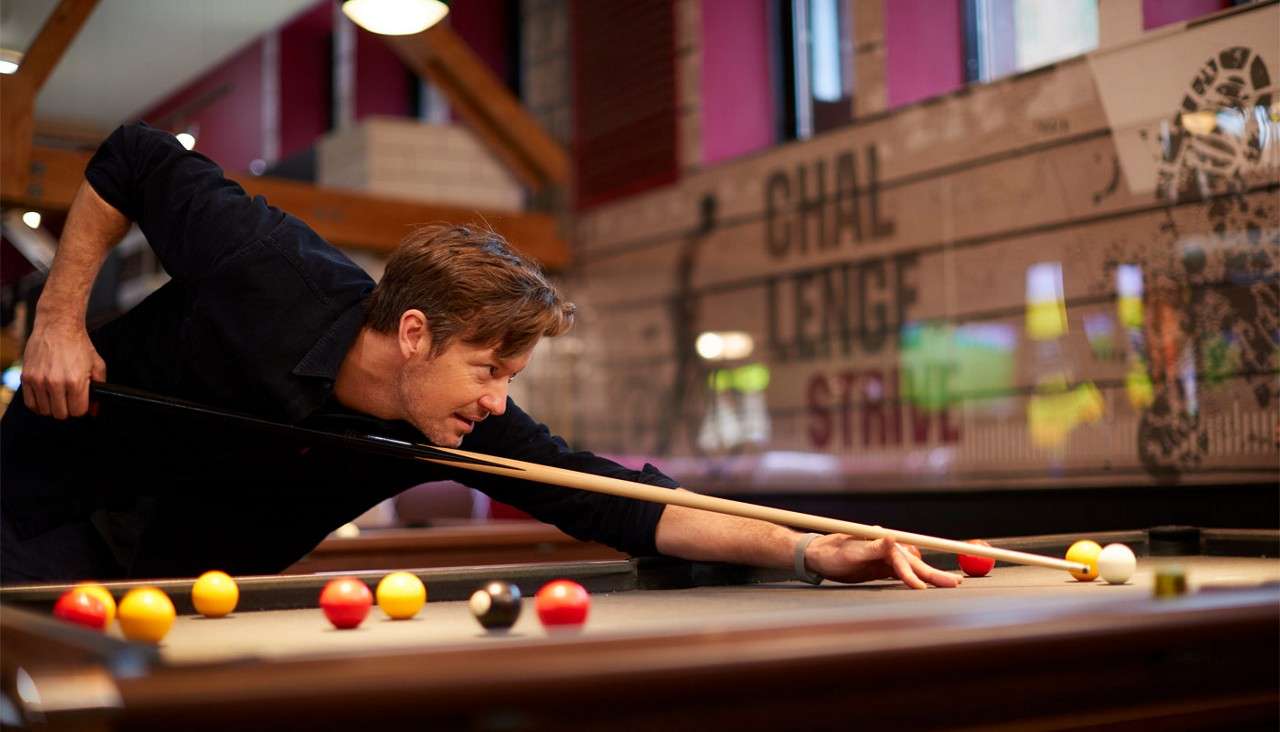 A person leans and aims a cue to strike the white ball on a pool table; red, yellow, and black balls nearby; casual bar setting; wall text: CHAL LENGE STRIVE.