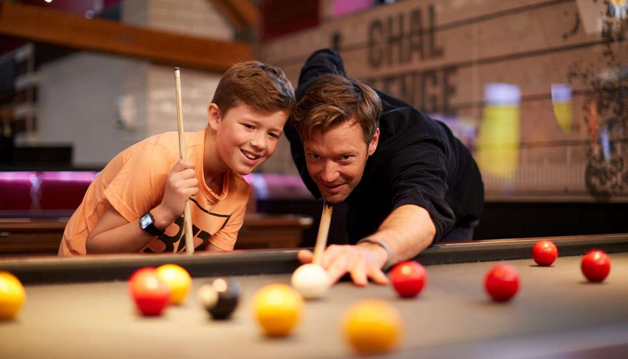 Man aims cue at white ball as smiling boy watches, playing pool on a table with red and yellow balls in a bar. Visible text: “CHAL LENGE”.