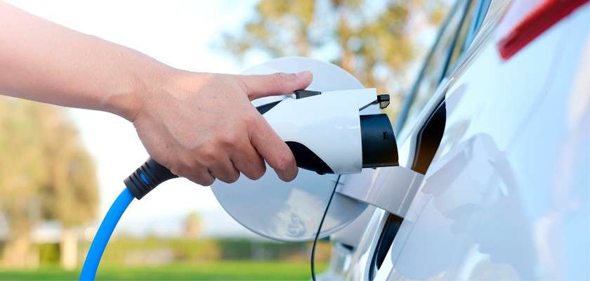 Charging plug connects to a white electric car's port, a hand inserting the blue-cabled connector outdoors with blurred trees and sky.