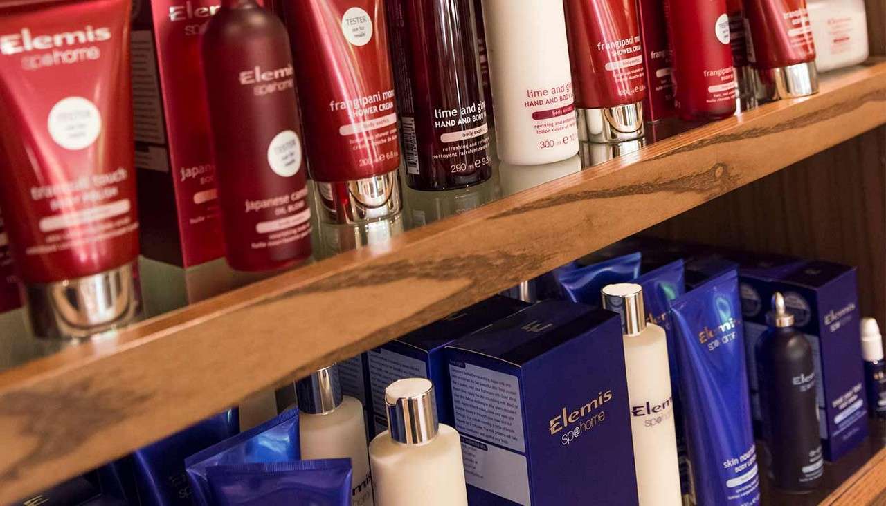Skincare bottles and tubes line wooden shelves in a store display. Text: Elemis, Sp@home, TESTER, frangipani monoi SHOWER CREAM, Japanese Camellia Oil Blend, lime and ginger HAND AND BODY…, skin…, 290 ml e, 300 ml e.