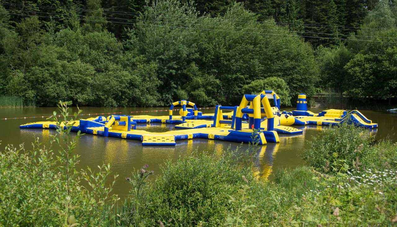 Inflatable water-park obstacles float quietly, featuring slides, tunnels, and climbing frames; they sit on a calm, greenish lake bordered by dense trees and shrubs under bright summer daylight.