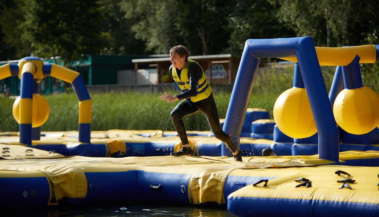 Person in a yellow life vest runs across a blue-and-yellow inflatable obstacle course, dodging large hanging balls, as water splashes around on a lakeside setup with reeds, trees, and buildings.