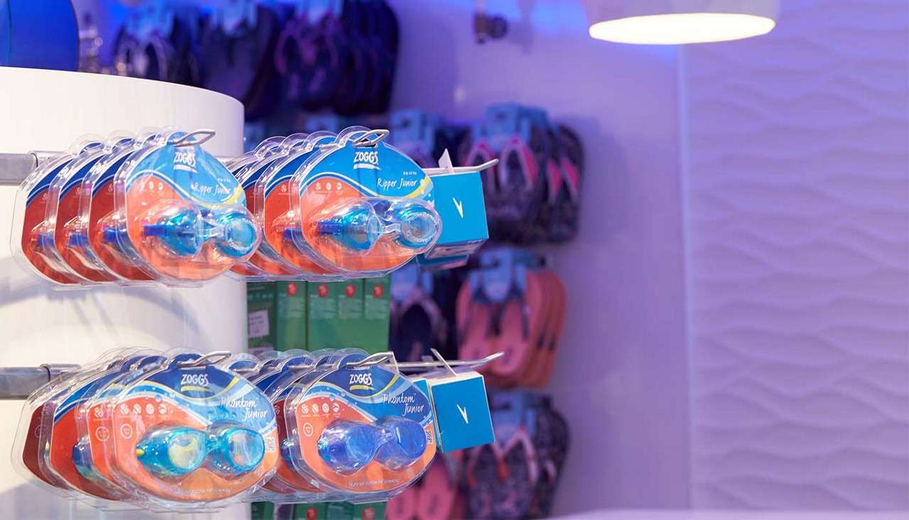 Packaged swimming goggles hang on display hooks for sale. Context: blue-lit retail area with a textured white wall and blurred flip-flops behind. Text: ZOGGS; Ripper Junior; Phantom Junior.