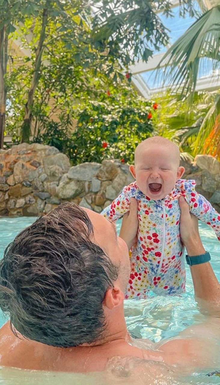 Baby in floral swimsuit is lifted above water by an adult, mouth wide open, in a shallow pool surrounded by rocks and lush tropical plants beneath a bright glass roof.