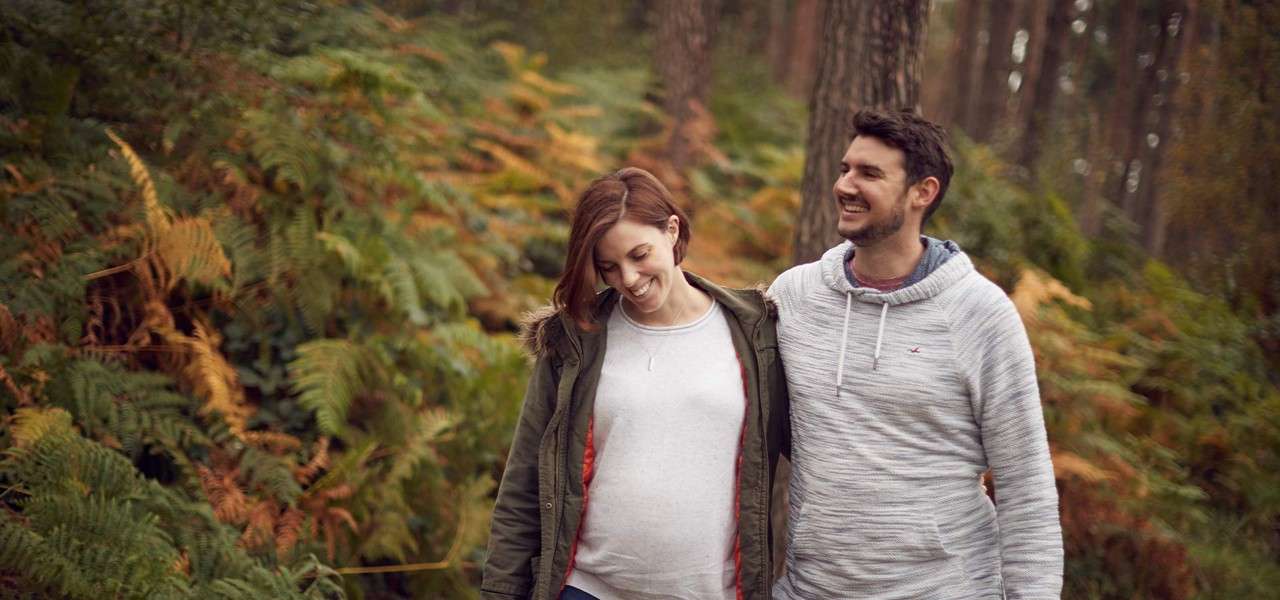 Two people walk together, smiling; one has an arm around the other’s shoulders, looking downward amid a forest with tall trees and dense green-and-brown ferns.