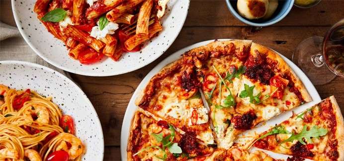 Pizza slices sit on a plate, topped with cheese, arugula, and sauce; nearby, bowls of rigatoni and shrimp spaghetti, plus small rolls, rest on a wooden table.