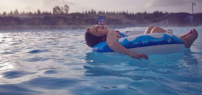 Child reclines laughing on a blue-and-white inner tube, floating on calm pool water; mist rises in cool evening light, with a rocky edge and treeline in the background.