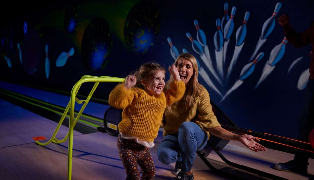 Child jumps and cheers, arms raised; adult kneels beside clapping. In a neon-lit bowling alley, a ball ramp sits by the lane, with pins mural glowing behind.