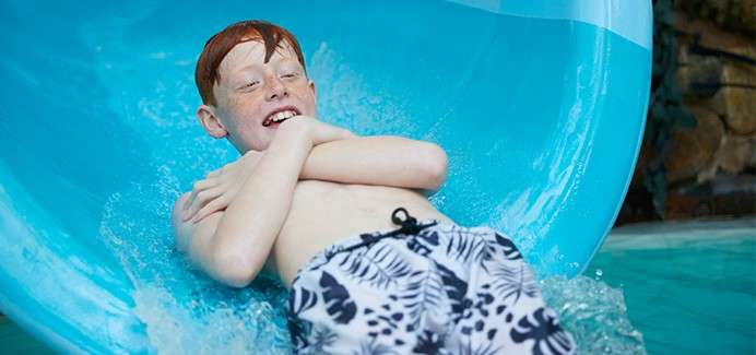 Boy slides down blue water slide, arms crossed over chest, smiling. Water splashes around him as he exits the tube into a pool area with a rock wall in the background.