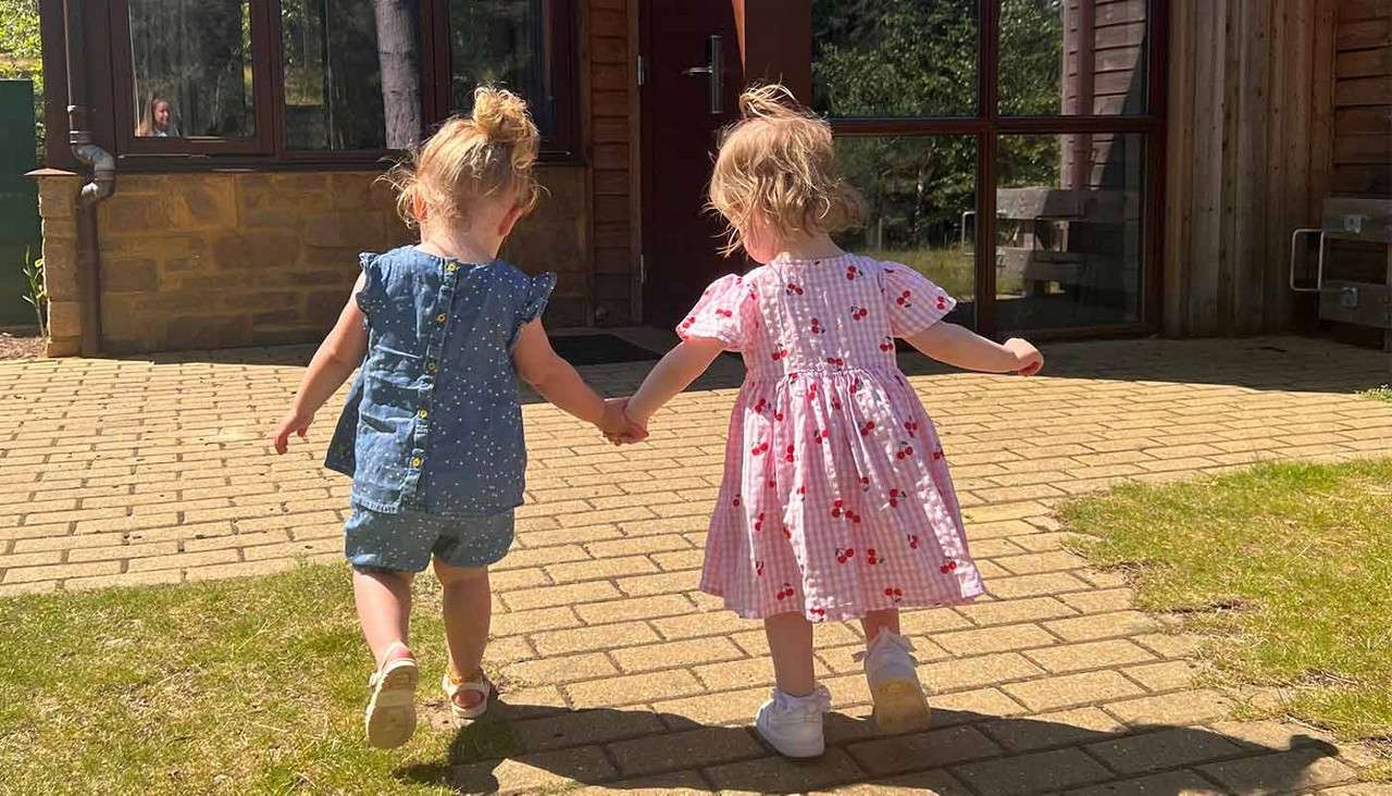 Two toddlers hold hands, walking away together on a sunlit brick path toward a wooden building with large windows; one wears a blue romper, the other a pink patterned dress.