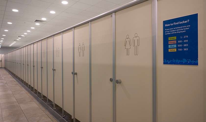 Row of beige locker-room stalls stands closed in a bright hallway; a wall sign provides instructions. Text: How to find locker? Press your wristband into one of our readers and the locker number will be displayed. Green 1–279; Orange 300–499; Blue 500–683; Yellow 700–983.