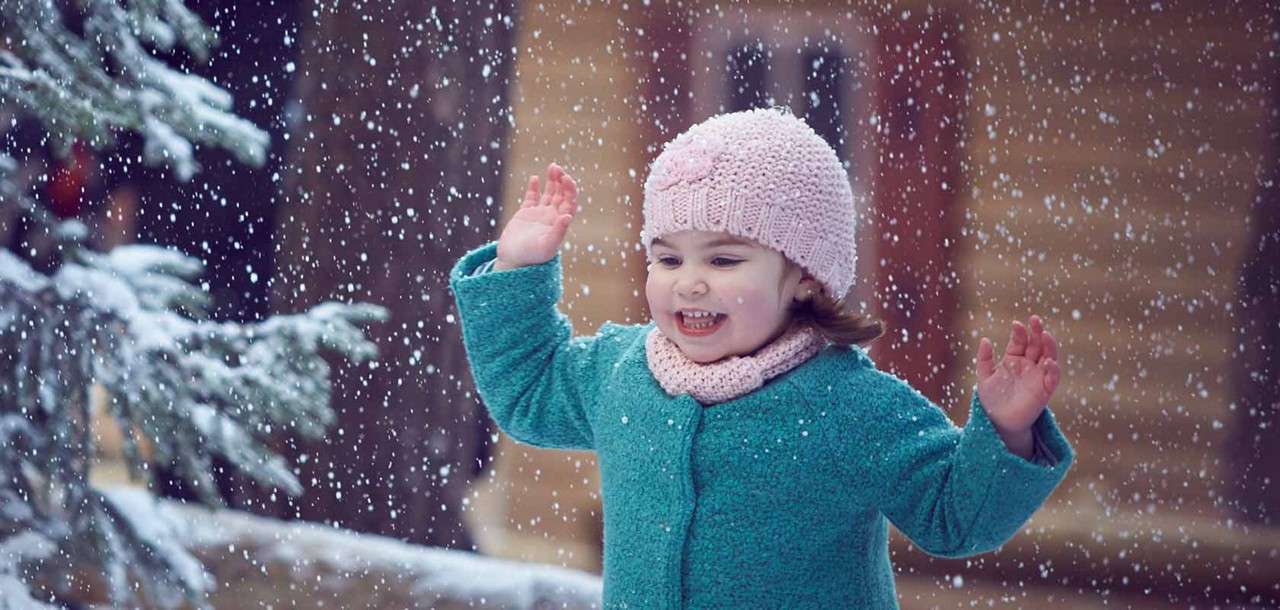 Child in teal coat and pink knit hat smiles with raised hands, catching falling snow; outdoors near a snow-dusted evergreen tree and blurred wooden buildings in soft winter light.