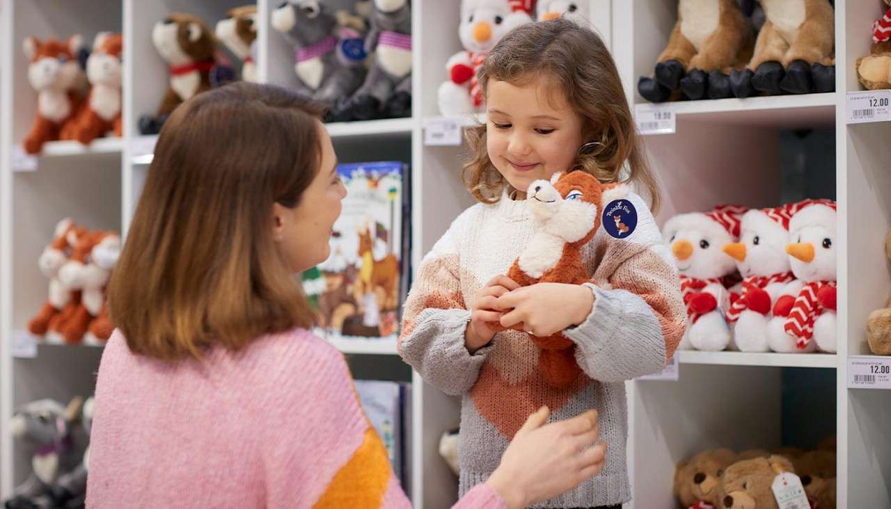 Girl hugs a fox plush while speaking with an adult; shelves of stuffed animals surround them in a toy store. Visible price labels read: 12.00.