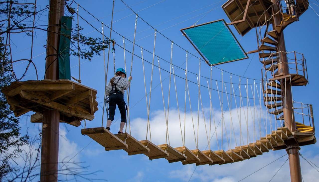 Harnessed participant crosses a wobbly wooden suspension bridge, gripping ropes. Elevated ropes course surrounds them with platforms, cables, and a spiral staircase, set outdoors against blue sky and scattered clouds.