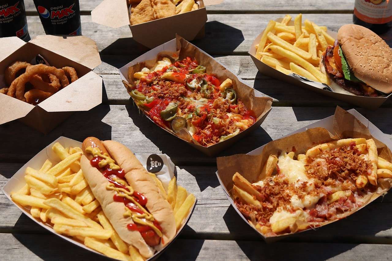 Assorted fast-food trays sit arranged on an outdoor picnic table: hot dog with ketchup and mustard, burger, onion rings, fries, loaded cheesy fries with jalapeños and bacon. Visible cans read: pepsi MAX, MAXIMUM TASTE NO SUGAR.