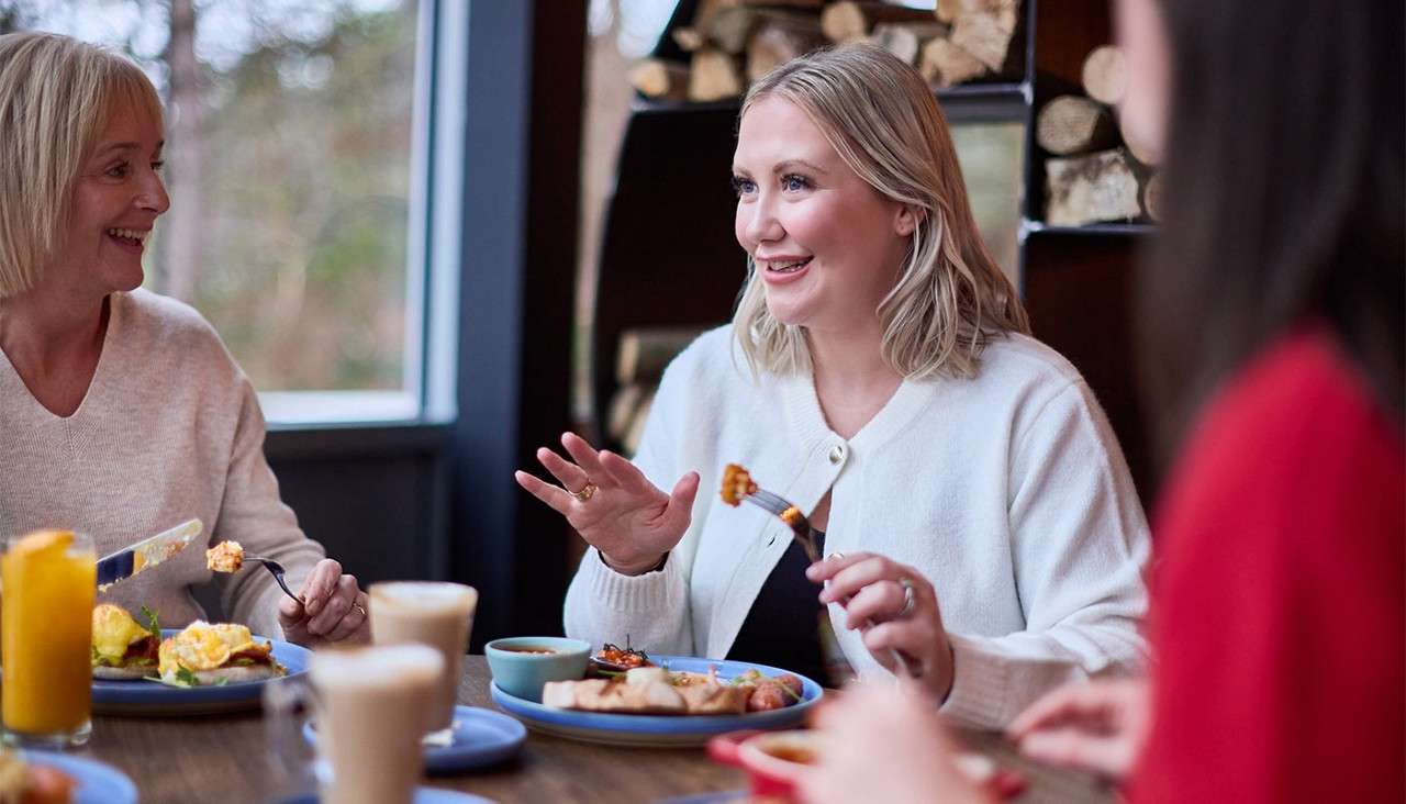 Woman holding a fork speaks animatedly while dining; companions eat nearby; plates of breakfast food, lattes, and juice on the table; cozy restaurant interior with a window and stacked firewood in the background.