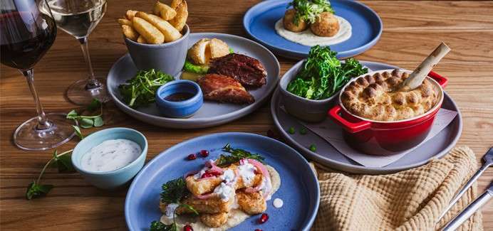 Assorted plated dishes sit arranged on a wooden table: meat, pies, fried bites, greens, dips, and chips, accompanied by two glasses of wine, cutlery, and a folded beige napkin.