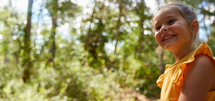 Child in a yellow sleeveless top smiles and looks right, head slightly tilted; sunlight filters through a leafy forest background, creating bright bokeh and a warm, outdoor atmosphere.