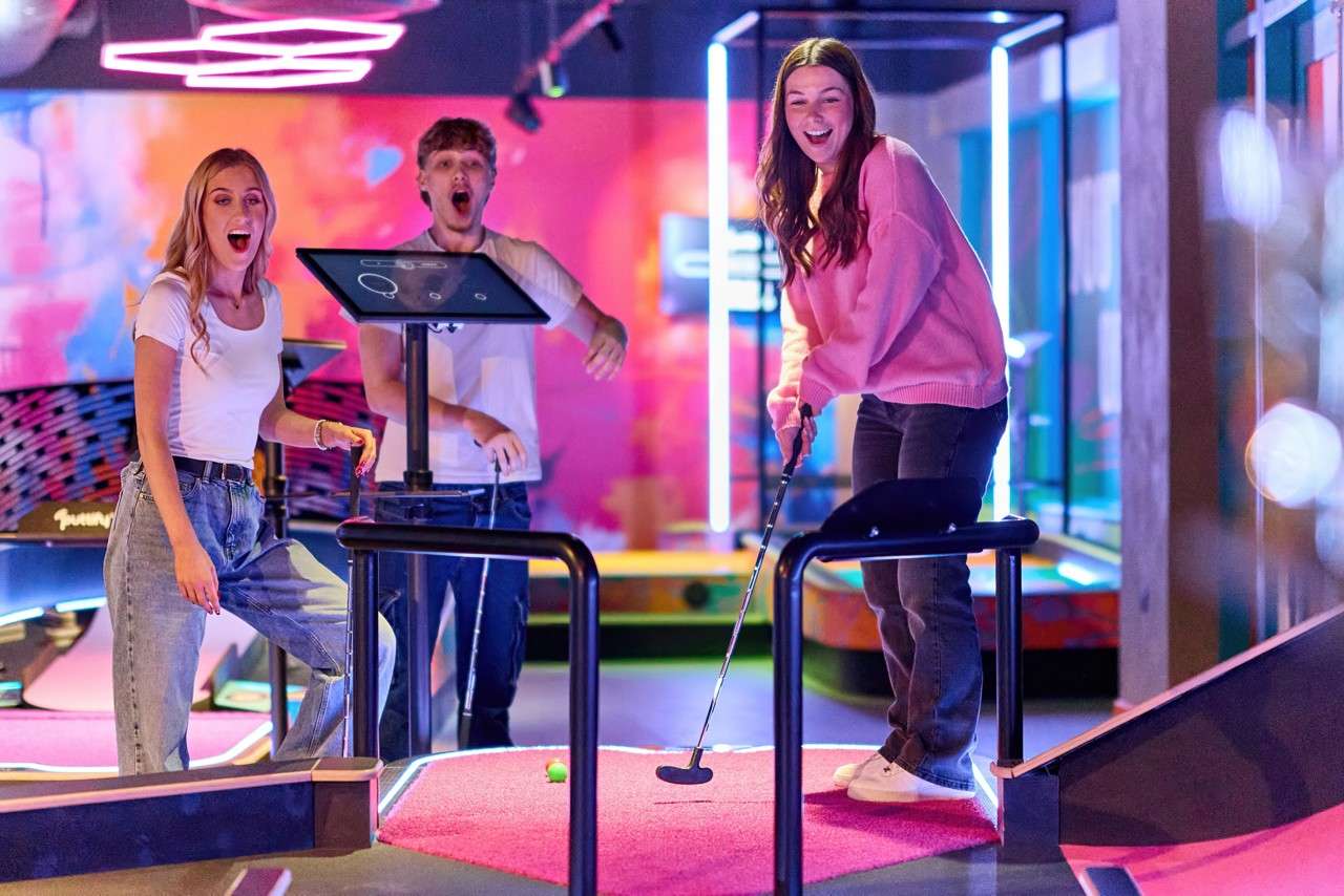Young woman putts a golf ball toward a hole, smiling. Two friends watch and cheer nearby in an indoor, neon-lit mini-golf course with a touchscreen kiosk and vibrant lights.