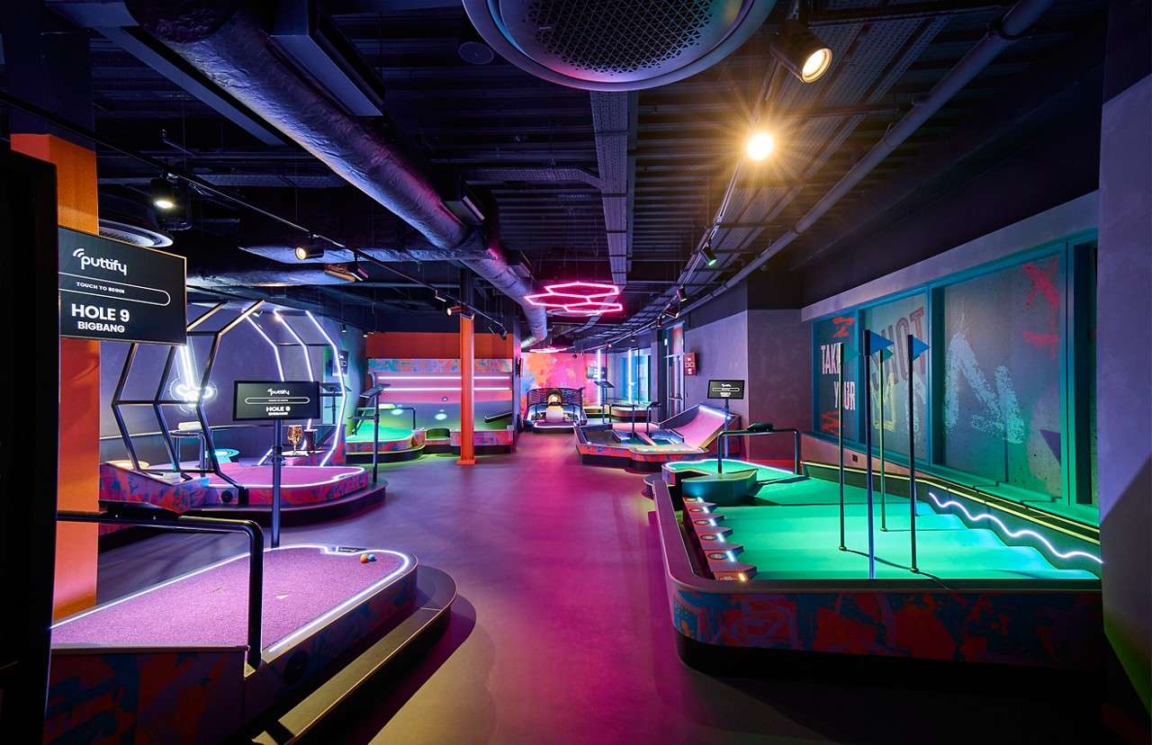 Mini-golf holes glow, guiding putters, inside a neon-lit indoor arcade with ramps, flags, and digital scoreboards.

Text: “puttify”, “TOUCH TO BEGIN”, “HOLE 9”, “BIGBANG”, “TAKE YOUR SHOT”.