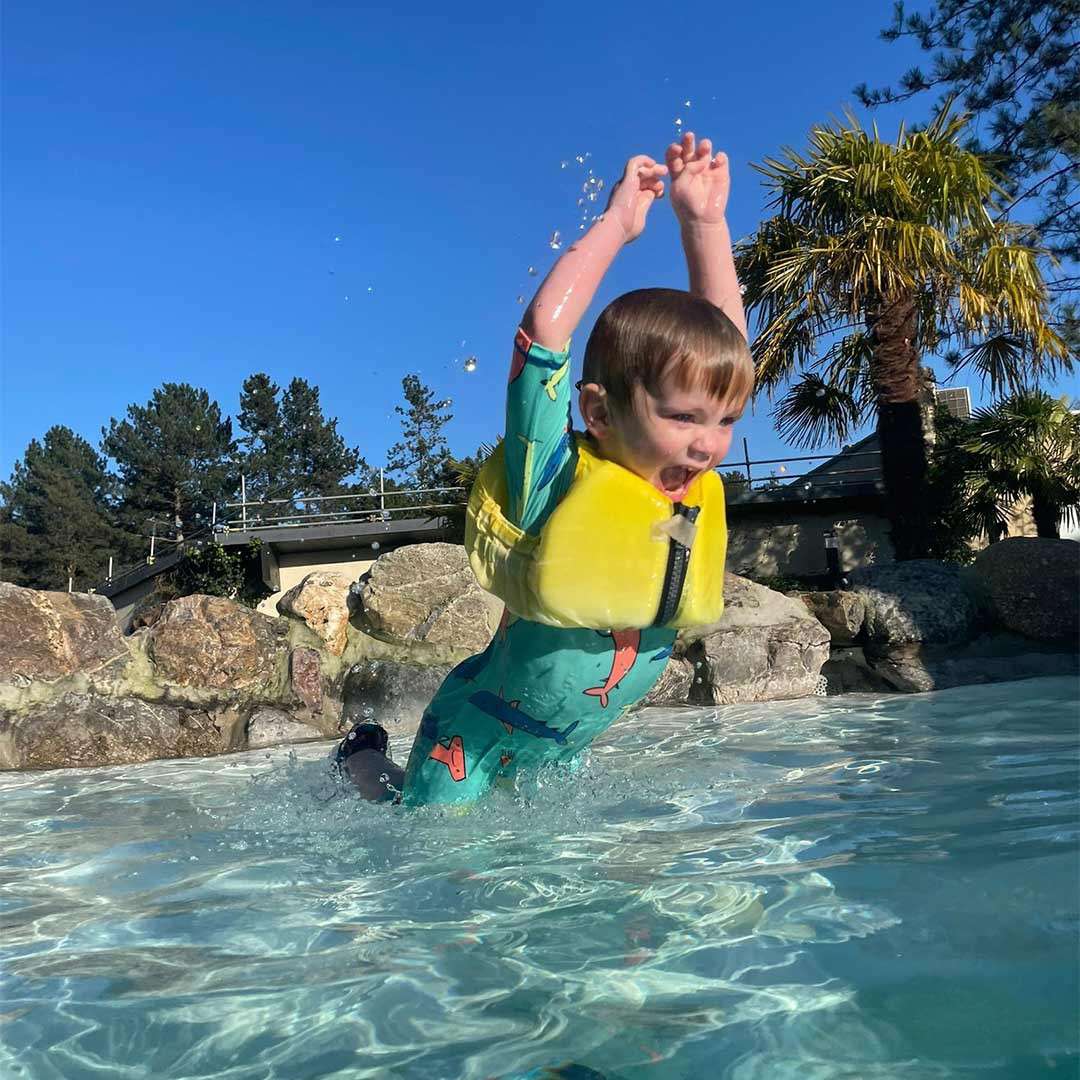 Child in a yellow life vest leaps into a shallow pool, arms raised, splashing water; outdoor setting with rock edging, palm trees, string lights, and a clear blue sky.