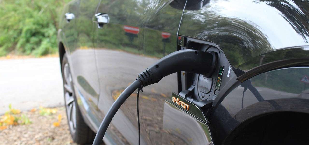 Electric car charges as a thick cable plugs into its side port, labeled e-tron; parked at a roadside pull-off with trees and a curving lane in the background.