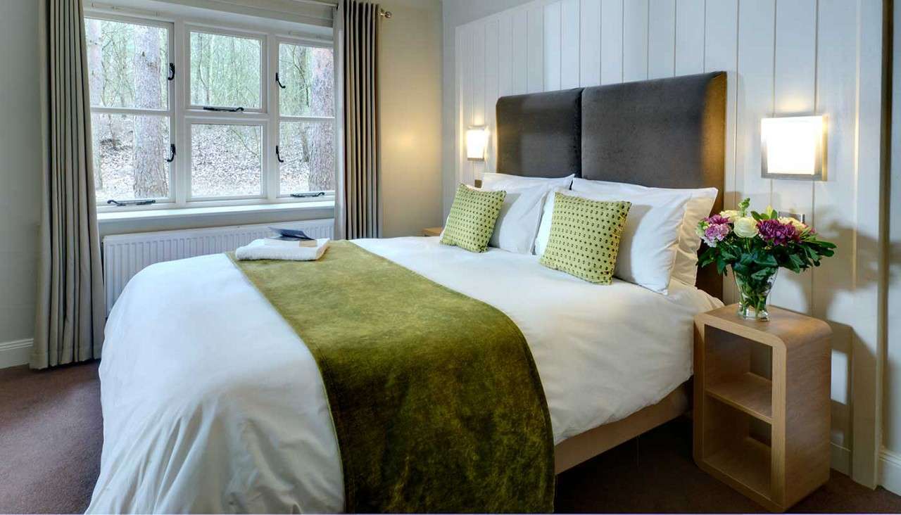 Bed, neatly made with white linens, green runner, and pillows, rests against a padded headboard; lamps glow as a curtained window shows woodland; bedside table holds flowers and towels.