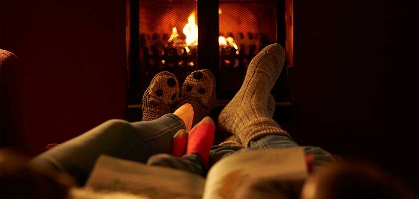 Socked feet rest, pointing toward a glowing fireplace; people lounge with legs outstretched, a blurred open book in the foreground, within a warm, dim room creating a cozy, relaxed atmosphere.