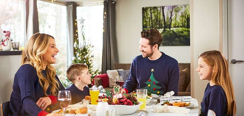Family of four—laughing—eat a festive meal at a dining table. Navy sweaters; one shows a Christmas tree. Candles, drinks, and decorations beside a window and lit tree.