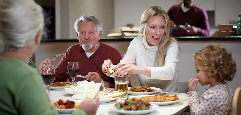 Family members eat and talk at a dining table, passing bread and food. Plates of pizza, salad, and drinks sit before them in a home kitchen, a person works behind.