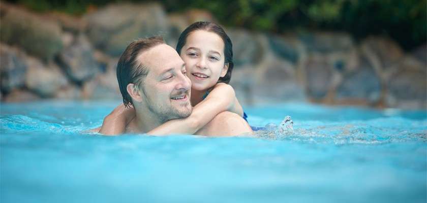 An adult swimmer carries a smiling child piggyback, gliding through a blue pool, with a stone edge and surrounding foliage in the background.