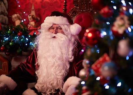 Santa Claus figure sits smiling, hands resting on lap, in a cozy, ornately decorated room with red wallpaper, glowing Christmas tree lights, ornaments, and festive garlands.